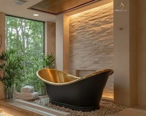 Luxurious bathroom with black and gold soaking tub surrounded by natural elements, stone wall, and large window overlooking green garden