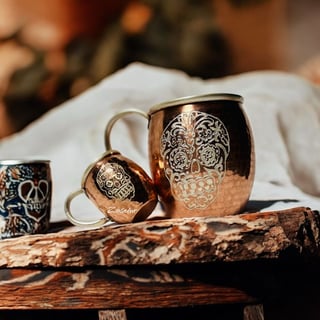 Copper mugs with ornate skull designs displayed on a rustic wooden surface with white cloth