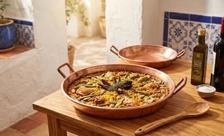 Paella in a large copper pan on a wooden table with olive oil and cooking utensils nearby